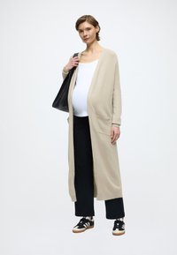 Beżowy dzianinowy kardigan z otwartym przodem i kieszeniami, noszony na białej bluzce i czarnych szerokich spodniach, zestawiony z czarno-kremowymi sneakersami.