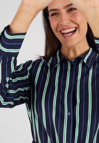 Blusa a righe blu navy con linee verticali verdi e blu, colletto e chiusura con bottoni. Tessuto liscio con maniche leggermente a sbuffo.