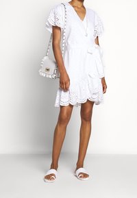 Abito bianco con scollo a V e maniche a sbuffo, caratterizzato da dettagli in occhielli, una cintura annodata e un orlo arricciato. Abbinato a sandali bianchi con cinturino.