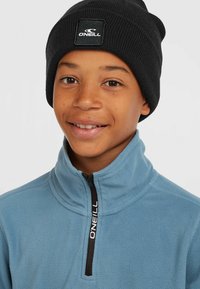 Light blue fleece jacket with a high collar and black zipper, featuring a logo on the zipper pull. Black ribbed beanie on head.