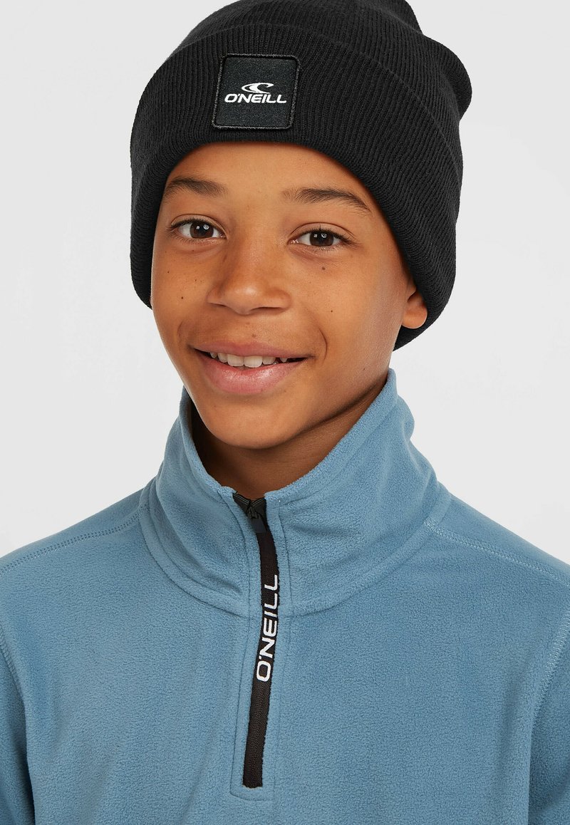 Light blue fleece jacket with a high collar and black zipper, featuring a logo on the zipper pull. Black ribbed beanie on head.