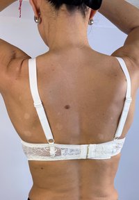 Sostenedor de encaje blanco en la espalda con tirantes ajustables, que presenta un cierre de gancho y ojo y un diseño de encaje texturizado. Tono de piel visible en la parte posterior.
