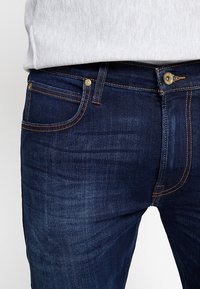 Gros plan d'une personne portant un jean en denim bleu foncé avec des coutures visibles et un bouton en métal, associé à une chemise gris clair.