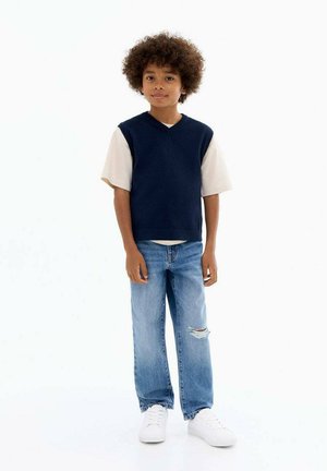 Gilet sans manches bleu marine en tricot sur un T-shirt crème, associé à un jean déchiré bleu clair et des baskets blanches. Texture lisse, coupe décontractée.