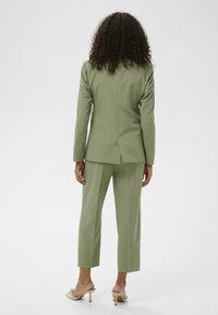 Costume vert clair sur mesure avec un blazer ajusté et un pantalon court, présentant une texture lisse et une fente à l'arrière. Porté avec des talons beiges.