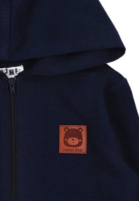 Felpa con zip color navy e un grande cappuccio. Presenta una toppa in pelle marrone con un grafiche di un orso e il testo "Sweet Bear". Tessuto morbido.