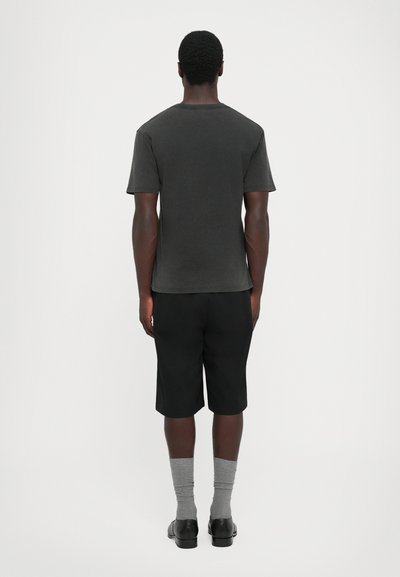 Adulto de costas, vestindo uma camisa de manga curta cinza escura, calções pretos, meias cinzas e sapatos pretos, em frente a um fundo simples.