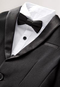 Veste de smoking noire avec revers en satin, chemise blanche et nœud papillon noir sur un fond uni clair.