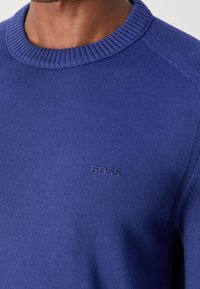 Gros plan d'une personne portant un pull en maille bleu avec un col rond côtelé et le logo "BOSS" brodé sur la poitrine.