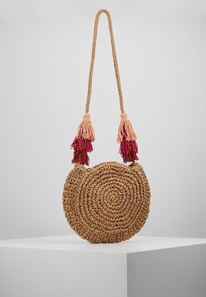 Sac bandoulière rond en paille tissée avec une lanière tressée et des pompons rouges et roses suspendus des deux côtés.