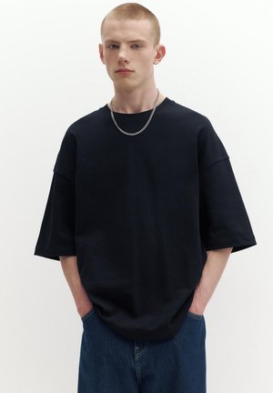 Bărbat tânăr cu păr scurt, purtând un tricou negru supradimensionat, un colier argintiu și blugi albaștri, cu mâinile în buzunare.