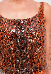 Close-up van een rood en zwart pailletten jurk met metalen knoopdetails en een getatoeëerde arm zichtbaar aan de rechterzijde.