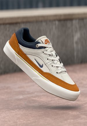 Nike SB sneaker i hvid, rustorange og marineblå, der svæver over en lyserød betonoverflade med en grå sløret baggrund.