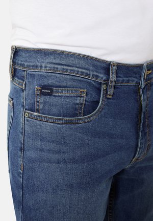 Denim-Jeans in Mittelblau mit figurnahen Schnitt, Seitentaschen und einer Gesäßtasche mit Markenlabel. Strukturierten Stoff mit sichtbaren Nähten.