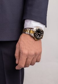 Montre-bracelet en métal doré et argenté avec un cadran noir, présentant des index des heures argentés et une fenêtre de date agrandie, portée sur une main en costume.