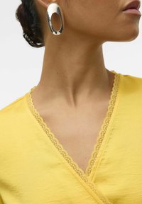 Žlutý bavlněný top s krajkovým výstřihem ve tvaru vlnky. Stříbrné oválné náušnice zdůrazňují límec. Viditelná hladká textura a jemný materiál.