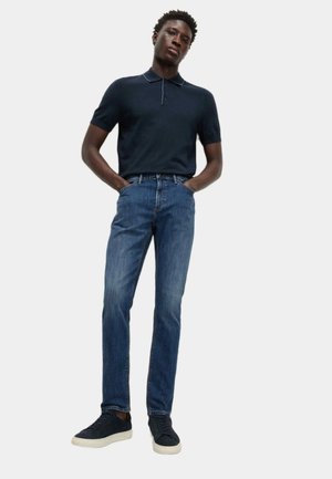 Mand står med hænderne i lommerne iført en mørkeblå polotrøje, blå jeans og sorte sneakers på en hvid baggrund.