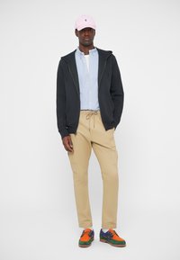 Μαύρο φούτερ με φερμουάρ πάνω από ανοιχτό μπλε πουκάμισο, χακί παντελόνι cargo και πολύχρωμα αθλητικά παπούτσια με ροζ καπέλο. Καθημερινή εμφάνιση με ορατές τσέπες.