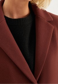 Ruskea villasekoituspäällystakki lovikauluksella, yhdistetty mustaan ribbineulepuseroon. Takissa on pehmeä, teksturoitu pinta.