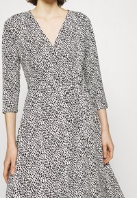 Wickelt Kleid aus hellbeigem Stoff mit schwarz gesprenkeltem Muster, tiefem V-Ausschnitt, Dreiviertelärmeln und einem Bindeband in der Taille.