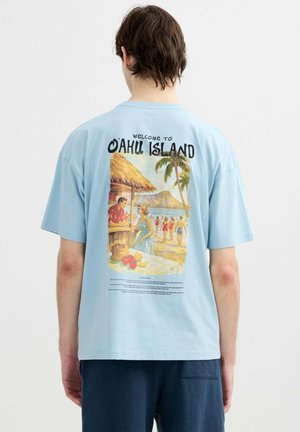 Persona que lleva una camiseta azul clara con una escena vintage de la playa de la isla de Oahu y palmeras impresas en la espalda.
