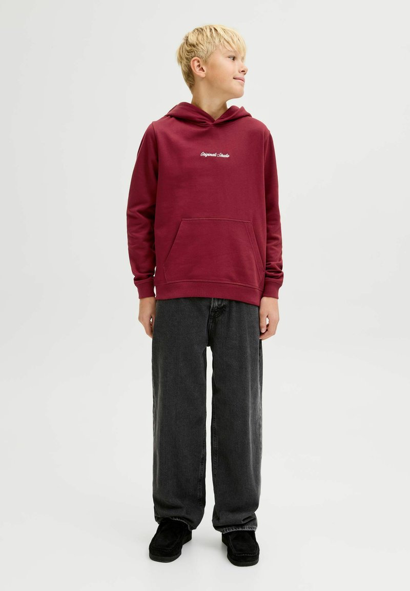 Sudadera marrón con bolsillo canguro, con texto blanco en el pecho. Combinada con pantalones de mezclilla negros holgados y zapatos negros.