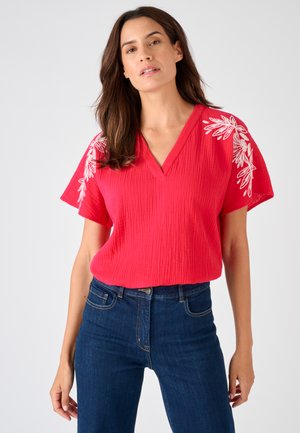 Mujer que lleva una blusa roja texturizada con escote en V y bordado floral blanco en las mangas, y vaqueros azul oscuro, de pie contra un fondo liso.
