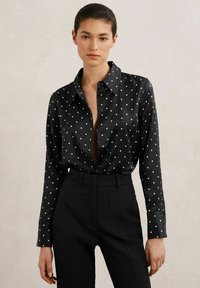 Mustast satiinist pikkade varrukatega pluus valgete täppidega, ees nööbitav ja kraega. Sobitatud kõrge vöökohaga mustade pükstega.