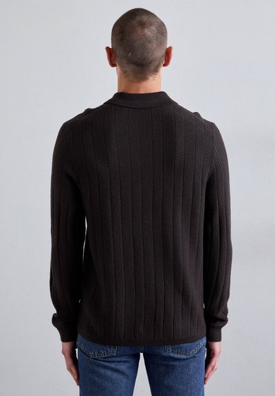 Temno rjav pleten pulover z navpičnimi rebrastimi detajli, dolgimi rokavi in visokim ovratnikom, v kombinaciji z modrimi kavbojkami. Očitna tekstura tkanine.
