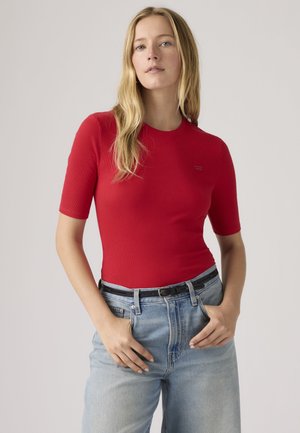 Giovane donna con capelli biondi lunghi che indossa una maglietta aderente rossa a maniche corte e jeans azzurri a vita alta con cintura nera.