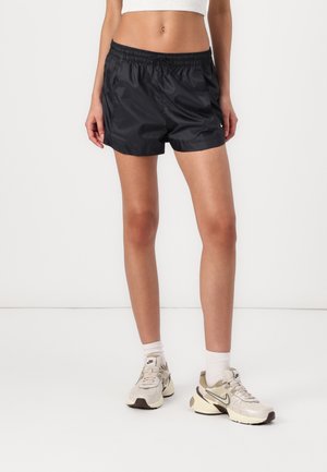 Kobieta ubrana w czarne sportowe szorty, białe skarpetki oraz beżowe buty Nike, stojąca na tle jednolitego białego tła.