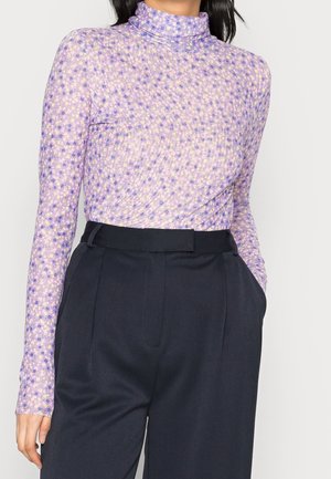 Mujer con un jersey de cuello alto floral de manga larga en color lavanda metido dentro de unos pantalones negros a medida de talle alto con las manos en los bolsillos.