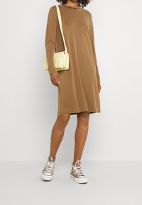 Brun, långärmad klänning med en mjuk textur, tillsammans med en vadderad gul handväska och mönstrade sneakers. Avslappnad, ledig passform.