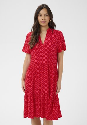 Kobieta w czerwonej sukience do kolan z krótkimi rękawami i geometrycznym diamentowym wzorem, stojąca na tle jednolitego jasnego tła.