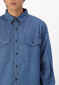 Camicia in denim di tonalità blu, con chiusura a bottoni, due tasche frontali e colletto. Presenta cuciture sottili e un'etichetta del marchio sulla tasca.