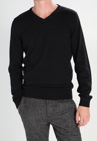 Pull noir à col en V en maille douce et légère. Comprend des manches longues et un ourlet côtelé. Porté par-dessus un pantalon gris texturé.