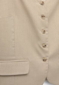 Tissu beige à fines rayures avec cinq boutons beiges et une poche à rabat sur le côté gauche, partie d'un vêtement sur mesure.