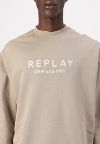 Bež dukserica s okruglim izrezom, s izvezenim natpisom "REPLAY DNM GDS 1981" u kontrastnoj boji. Mekani materijal, širok kroj i rebrasti ovratnik.