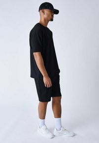 Chemise noire à manches courtes et shorts noirs, tous deux de coupe décontractée. Baskets blanches et chaussettes blanches jusqu'aux chevilles complètent le look. Casquette noire portée.