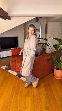 Lysegrå badekåbe i blødt materiale med bælte, båret over grå bukser og beige sko. Indendørs omgivelser med en plante og møbler.