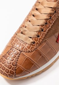 Bruna sneakers med krokodilsmönster, satinbeiga skosnören, mockadetaljer och vit sula, som visar upp detaljer i strukturerat läder.