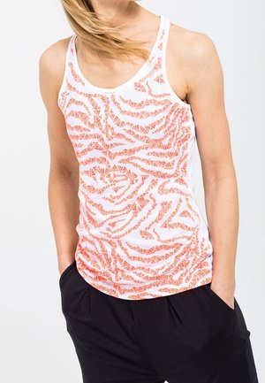 Personne portant un débardeur blanc avec un motif abstrait orange, associé à un pantalon noir, debout les mains dans les poches.