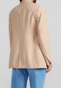 Béžový blazer s hladkým materiálem, strukturovaným střihem, jedním rozparkem na zadní části a dlouhými rukávy, zkombinovaný s světle modrými džíny.