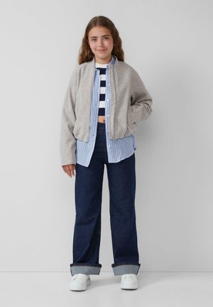 Grijze bomberjack, gestreept blauw-wit shirt eronder, donkerblauwe wijde jeans met opgerolde zoom, en witte sneakers.
