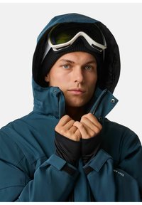 Türkise isolierte Jacke mit Kapuze, durchgehendem Reißverschluss, schwarzen Bündchen und hohem Kragen. Weißer Skibrille und eine schwarze Mütze werden getragen.
