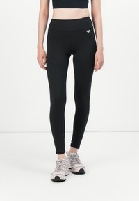 Kvinne iført svarte høytaljede leggings med en liten logo på hoften og lysegrå joggesko, stående mot en hvit bakgrunn.