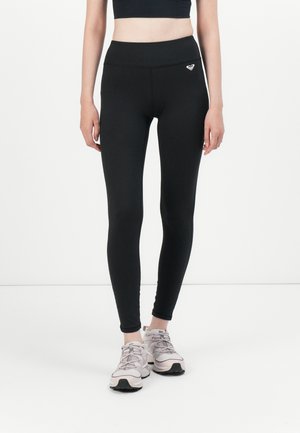 Femme portant des leggings taille haute noirs avec un petit logo sur la hanche et des chaussures de course gris clair, debout contre un fond blanc.