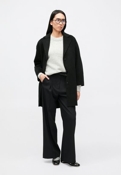 Nadrozmerný čierny kabát cez sivý pletený sveter, doplnený čiernymi nohavicami s prúžkami a lesklými čiernymi topánkami, so zaoblenými modrými okuliarmi.