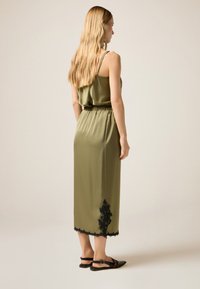 Abito in satin verde oliva con spalline sottili regolabili, fascia elastica in vita e dettagli in pizzo nero all'orlo. Sandali neri piatti completano il look.