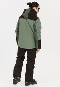 Wasserdichte Skijacke in Grün und Schwarz, mit Kapuze, lässigem Schnitt und strapazierfähigem Stoff. Kombiniert mit schwarzen Hosen und Skischuhen.